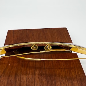 Puede incluir: Gafas de sol de metal en tono dorado con patillas de alambre fino y almohadillas nasales ajustables. El puente est&aacute; grabado con el texto B&L RAY-BAN U.S.A. Las lentes tienen un tono &aacute;mbar marr&oacute;n c&aacute;lido, mostrando la estructura de la montura.