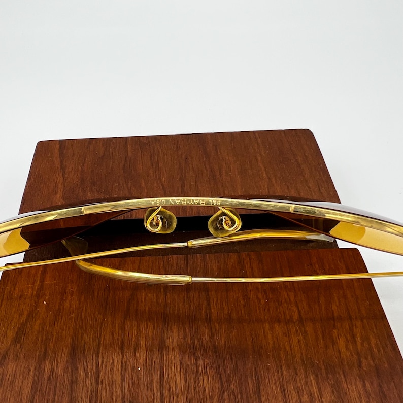 Puede incluir: Gafas de sol de metal en tono dorado con patillas de alambre fino y almohadillas nasales ajustables. El puente est&aacute; grabado con el texto B&L RAY-BAN U.S.A. Las lentes tienen un tono &aacute;mbar marr&oacute;n c&aacute;lido, mostrando la estructura de la montura.