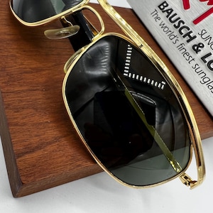 Puede incluir: Gafas de sol con montura dorada y lentes oscuras, apoyadas sobre una superficie de madera. Las gafas de sol tienen un dise&ntilde;o cl&aacute;sico de aviador. El texto "BAUSCH & LOMB" es visible en el fondo.