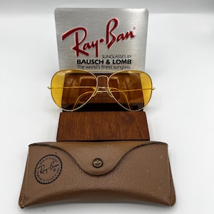 Puede incluir: Gafas de sol tipo aviador en tono dorado con lentes amarillas, situadas frente a un letrero expositor plateado de Ray-Ban Bausch & Lomb. Un estuche de cuero marr&oacute;n texturizado con cierre de bot&oacute;n y el logo de Ray-Ban est&aacute; en primer plano.