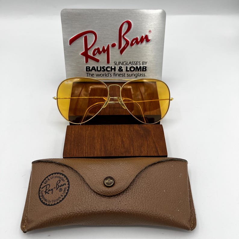 Puede incluir: Gafas de sol tipo aviador en tono dorado con lentes amarillas, situadas frente a un letrero expositor plateado de Ray-Ban Bausch & Lomb. Un estuche de cuero marr&oacute;n texturizado con cierre de bot&oacute;n y el logo de Ray-Ban est&aacute; en primer plano.