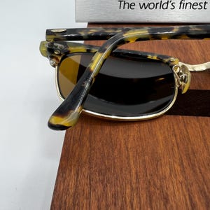 May include: Close-up of sunglasses with a gold frame and dark lenses. The temples are a tortoiseshell pattern. The text "The world's finest" is visible in the background.