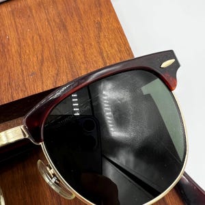May include: Vintage sunglasses with a dark brown tortoiseshell frame and gold-tone accents. The lenses are dark, and the frame has a classic, retro design. The text "BAUSCH & LOMB RAY" is visible on the frame.