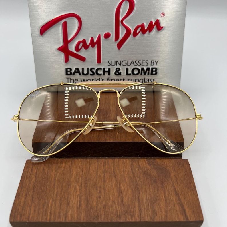 May include: Gold aviator sunglasses with brown gradient lenses. The sunglasses are on a wooden display stand with a Ray-Ban box behind them. The box has the text "Ray-Ban Sunglasses by Bausch & Lomb The world's finest sunglass" on it.