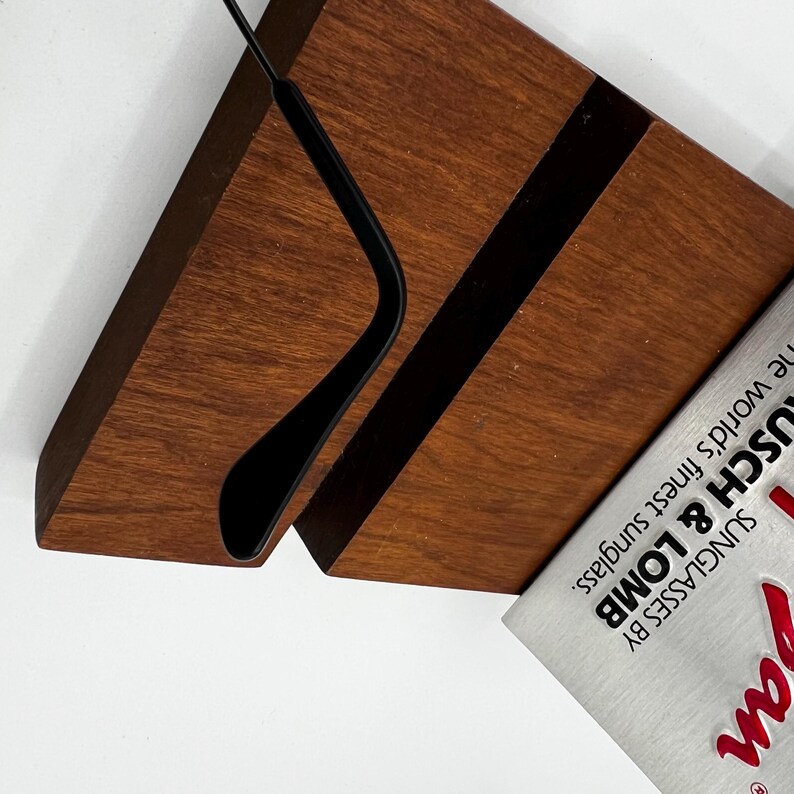 K&ouml;nnte beinhalten: Ein Sonnenbrillen-Displayst&auml;nder aus Holz mit dunkelbraunem Finish. Der St&auml;nder hat einen schwarzen Einsatz und einen gebogenen schwarzen Metallarm zum Halten der Sonnenbrillen. Ein Magazin mit dem Text "Bausch & Lomb Sunglasses" ist sichtbar.