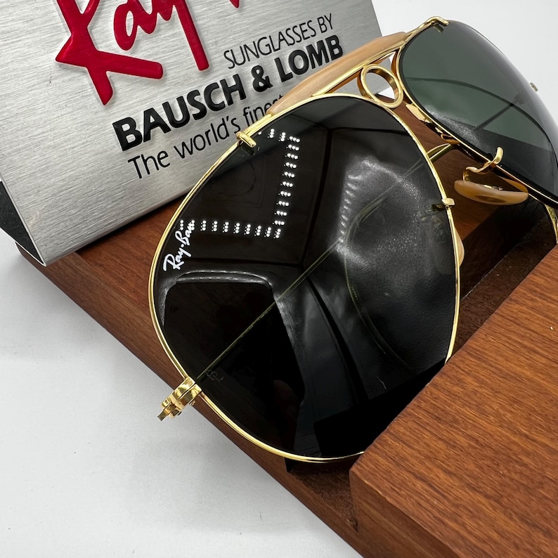 Pode incluir: &Oacute;culos de sol aviador com arma&ccedil;&atilde;o dourada e lentes escuras. O logotipo da Ray-Ban &eacute; vis&iacute;vel na lente. Os &oacute;culos de sol s&atilde;o exibidos em um suporte de madeira, com uma placa de prata que diz "SUNGLASSES BY BAUSCH & LOMB The world's finest."