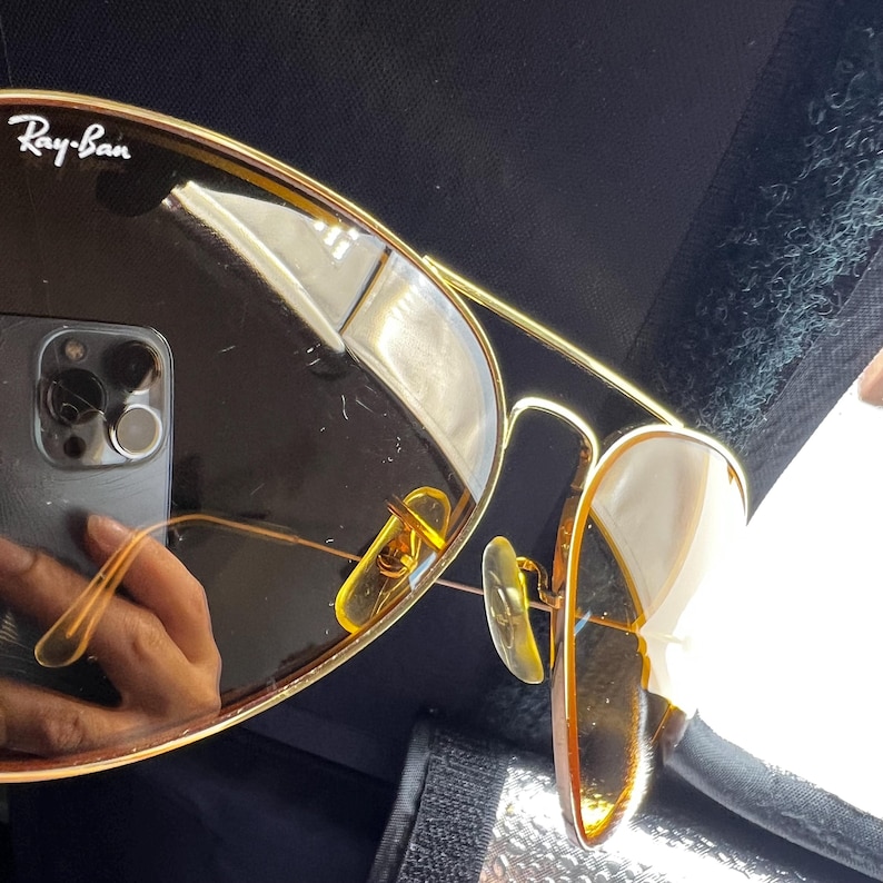 Puede incluir: Gafas de sol con montura de metal en tono dorado y lentes tintadas en marr&oacute;n. La lente izquierda presenta el logotipo blanco de Ray-Ban en la esquina superior. Se aprecian las varillas finas de alambre y las almohadillas nasales ajustables.