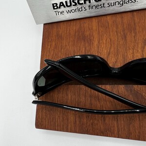 May include: Black sunglasses with a glossy finish are displayed on a wooden surface. The text "BAUSCH & The world's finest sunglass" is visible in the background. The sunglasses have a classic design.