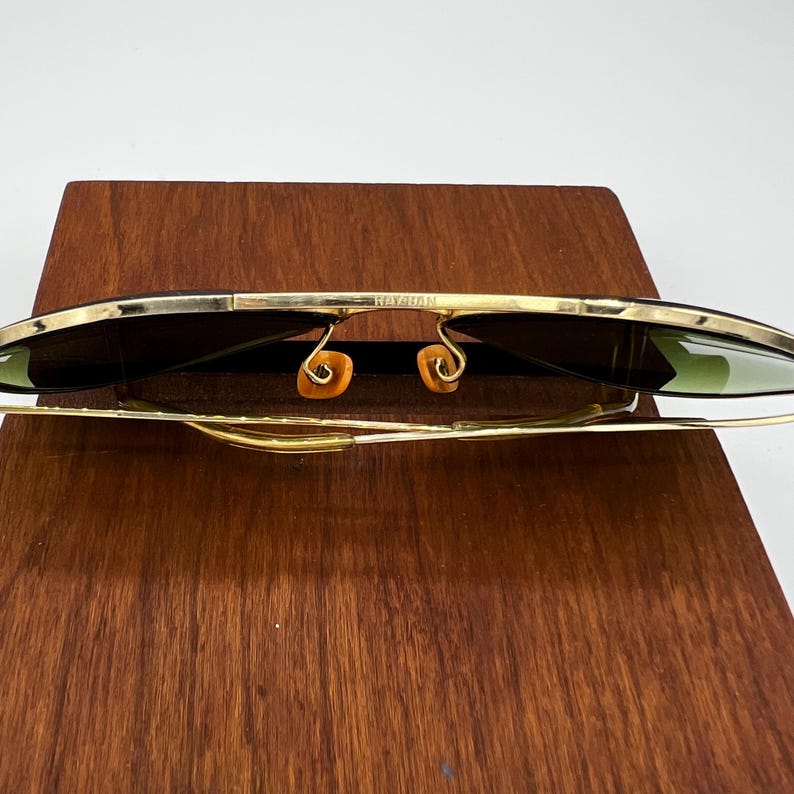 May include: Gold-rimmed sunglasses with dark lenses. The sunglasses have a classic design with a thin gold frame and a bridge. The lenses are dark, providing shade and protection from the sun. The sunglasses are displayed on a wooden surface.