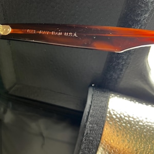 Puede incluir: Primer plano de unas gafas de sol Ray-Ban con montura carey. El texto "B&L RAY-BAN U.S.A." es visible en la patilla. Las gafas de sol est&aacute;n dentro de un estuche negro con interior dorado.