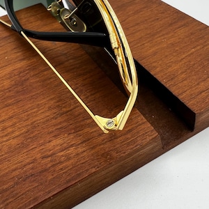 Puede incluir: Primer plano de gafas de sol con montura dorada y monturas negras. Las gafas descansan sobre una superficie de madera marr&oacute;n oscuro. Los detalles dorados y las bisagras son visibles, mostrando los detalles del dise&ntilde;o.