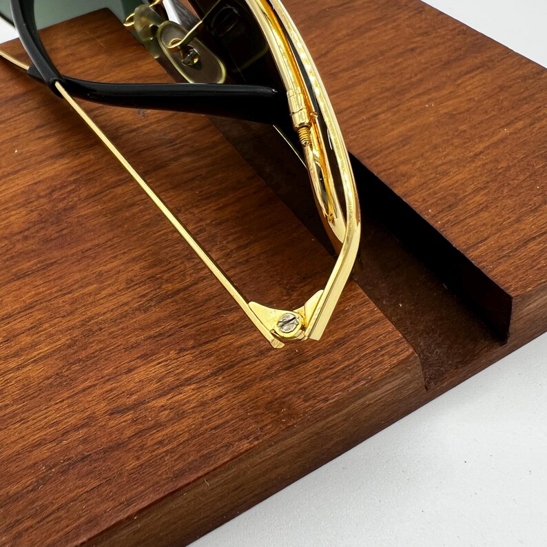 Puede incluir: Primer plano de gafas de sol con montura dorada y monturas negras. Las gafas descansan sobre una superficie de madera marr&oacute;n oscuro. Los detalles dorados y las bisagras son visibles, mostrando los detalles del dise&ntilde;o.