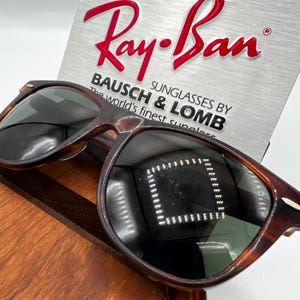 May include: Ray-Ban sunglasses with a tortoiseshell frame. The lenses are dark, reflecting the surrounding environment. The Ray-Ban logo is in red, above the text "BAUSCH & LOMB SUNGLASSES BY The world's finest sunglasses."