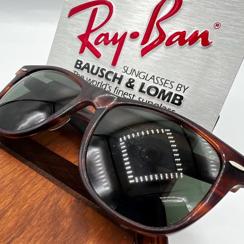 May include: Ray-Ban sunglasses with a tortoiseshell frame. The lenses are dark, reflecting the surrounding environment. The Ray-Ban logo is in red, above the text "BAUSCH & LOMB SUNGLASSES BY The world's finest sunglasses."