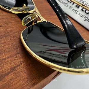 Puede incluir: Primer plano de gafas de sol con montura dorada y lentes negras. Las gafas de sol tienen un dise&ntilde;o elegante y moderno con una superficie reflectante. La imagen est&aacute; sobre un fondo de madera.