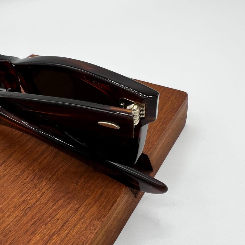 Puede incluir: Primer plano de gafas marrones con montura de carey sobre una superficie de madera. Las gafas tienen un dise&ntilde;o cl&aacute;sico con un acabado oscuro y brillante. Se ven las bisagras y las patillas.