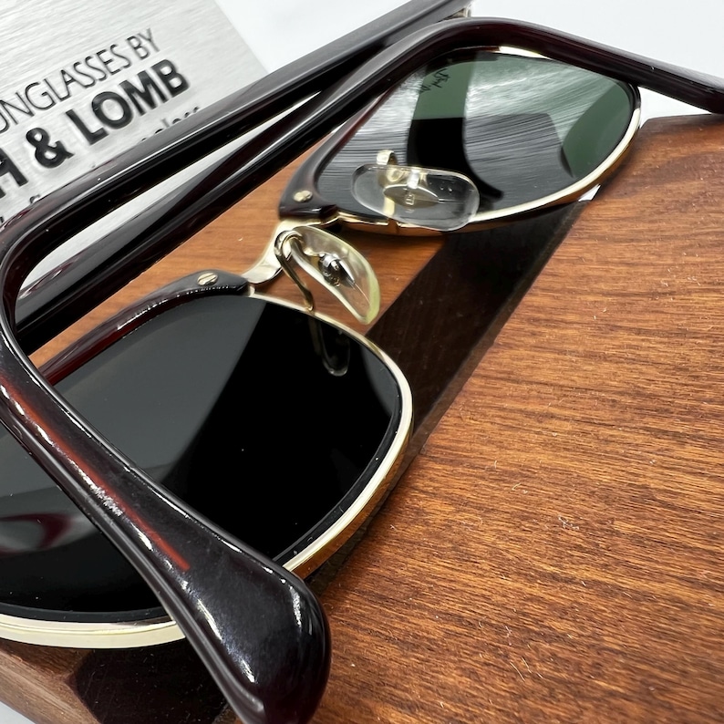 May include: Close-up of sunglasses with dark lenses and a gold frame. The frames are a dark brown color. The sunglasses are resting on a wooden surface.