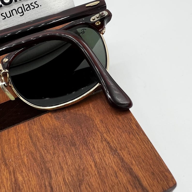 May include: Dark brown sunglasses with black lenses and gold-colored accents. The sunglasses are resting on a wooden surface. The text "sunglass" is partially visible in the upper left corner.