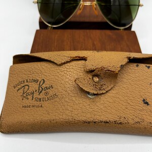 May include: Gold-framed aviator sunglasses with green lenses rest above a tan leather case. The case is embossed with "Ray-Ban Sunglasses" and "Made in U.S.A." The case has a worn, textured appearance.