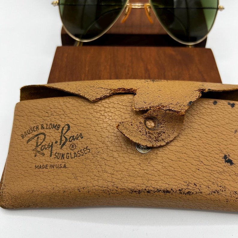 May include: Gold-framed aviator sunglasses with green lenses rest above a tan leather case. The case is embossed with "Ray-Ban Sunglasses" and "Made in U.S.A." The case has a worn, textured appearance.