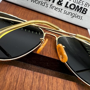 May include: Gold-framed sunglasses with dark lenses and amber nose pads. The sunglasses are resting on a wooden surface. The text "world's finest sunglass" is visible in the background.