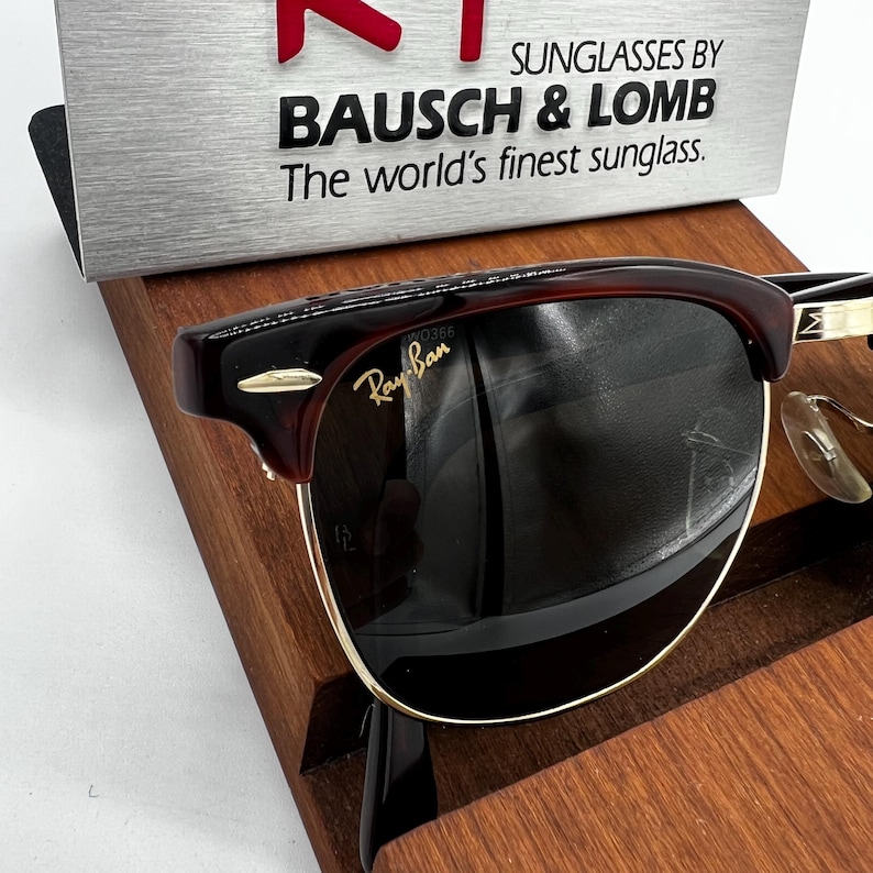 May include: Ray-Ban sunglasses with a dark burgundy frame and gold accents. The lenses are dark, and the brand name is visible on the lens. The sunglasses are displayed on a wooden stand, with the text "SUNGLASSES BY BAUSCH & LOMB" above.