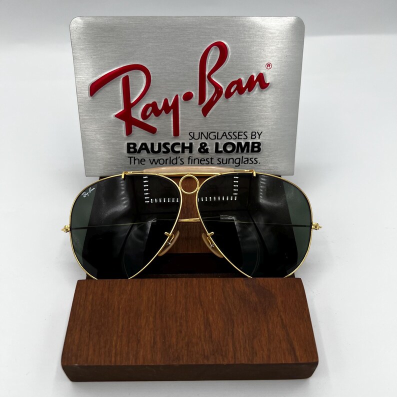 Pode incluir: &Oacute;culos de sol Ray-Ban com arma&ccedil;&atilde;o dourada e lentes escuras, apresentados em um estojo de madeira. Os &oacute;culos de sol est&atilde;o em frente a uma placa de prata com o logotipo da Ray-Ban e o texto: "SUNGLASSES BY BAUSCH & LOMB The world's finest sunglass."