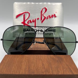 Pu&ograve; includere: Occhiali da sole aviator Ray-Ban neri con lenti verdi. Gli occhiali sono su una superficie di legno con una scatola argentata dietro. La scatola ha il logo Ray-Ban e il testo "BAUSCH & LOMB The world's finest sunglasses."