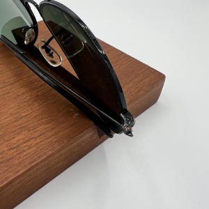 Pu&ograve; includere: Occhiali da sole aviator neri con lenti verdi piegati. Gli occhiali da sole sono appoggiati su una superficie di legno.