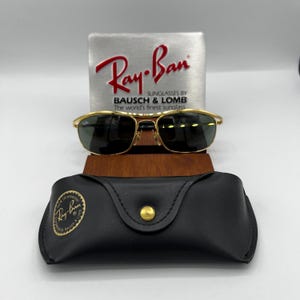 Puede incluir: Gafas de sol Ray-Ban con montura dorada y lentes oscuras, presentadas con un estuche de cuero negro y un letrero plateado con el logotipo de Ray-Ban. El letrero tambi&eacute;n incluye el texto "SUNGLASSES BY BAUSCH & LOMB The world's finest sunglass."