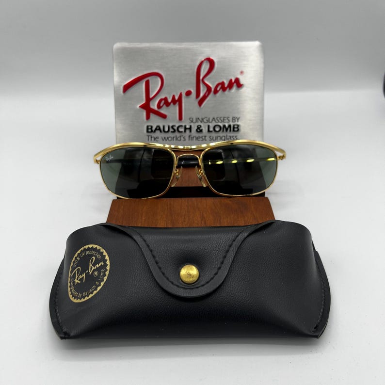 Puede incluir: Gafas de sol Ray-Ban con montura dorada y lentes oscuras, presentadas con un estuche de cuero negro y un letrero plateado con el logotipo de Ray-Ban. El letrero tambi&eacute;n incluye el texto "SUNGLASSES BY BAUSCH & LOMB The world's finest sunglass."