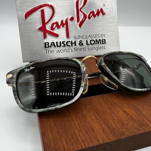Pu&ograve; includere: Occhiali da sole Ray-Ban con montatura verde e grigia marmorizzata, dettagli dorati e lenti scure. Il logo Ray-Ban &egrave; visibile sulla montatura. Un cartello argentato dietro gli occhiali recita "Ray-Ban" e "SUNGLASSES BY BAUSCH & LOMB".