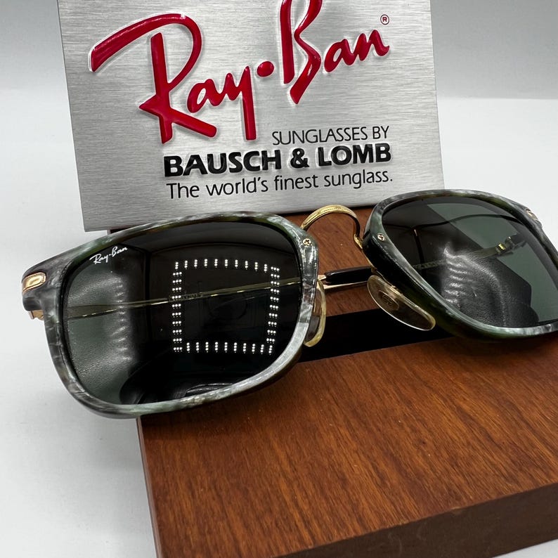 Pu&ograve; includere: Occhiali da sole Ray-Ban con montatura verde e grigia marmorizzata, dettagli dorati e lenti scure. Il logo Ray-Ban &egrave; visibile sulla montatura. Un cartello argentato dietro gli occhiali recita "Ray-Ban" e "SUNGLASSES BY BAUSCH & LOMB".
