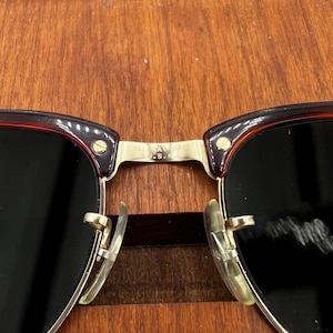 May include: Close-up of sunglasses with a dark brown frame and black lenses. The metal bridge and arms are gold-colored. The text "B&L USA" is visible on the bridge.