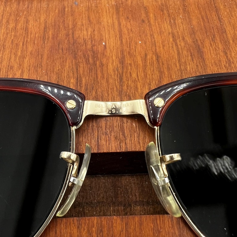 May include: Close-up of sunglasses with a dark brown frame and black lenses. The metal bridge and arms are gold-colored. The text "B&L USA" is visible on the bridge.