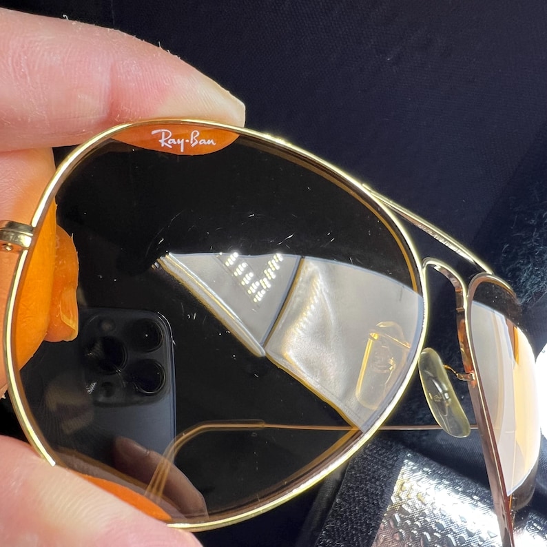 Puede incluir: Gafas de sol con montura de metal dorado y lentes oscuras. La lente superior derecha presenta el logotipo blanco de Ray-Ban impreso sobre un fondo naranja. La fina montura de alambre incluye almohadillas nasales ajustables y un puente cl&aacute;sico.