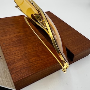 Puede incluir: Gafas de sol de metal en tono dorado con lentes tintadas en color &aacute;mbar. La montura tiene un acabado pulido fino con el logotipo de Ray-Ban visible impreso en la esquina superior del cristal derecho. Las varillas son delgadas y met&aacute;licas.