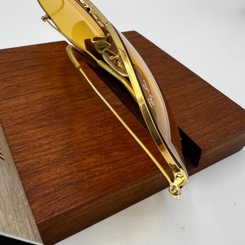 Puede incluir: Gafas de sol de metal en tono dorado con lentes tintadas en color &aacute;mbar. La montura tiene un acabado pulido fino con el logotipo de Ray-Ban visible impreso en la esquina superior del cristal derecho. Las varillas son delgadas y met&aacute;licas.