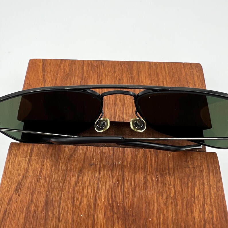 K&ouml;nnte beinhalten: Schwarze Aviator-Sonnenbrille mit dunklen Gl&auml;sern und einem d&uuml;nnen Metallrahmen. Die Sonnenbrille hat einen Doppelsteg und ein klassisches Design. Die Gl&auml;ser scheinen gr&uuml;n get&ouml;nt zu sein. Die Sonnenbrille liegt auf einer Holzoberfl&auml;che.