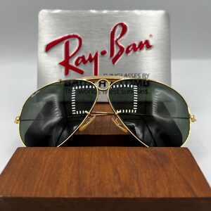 Pode incluir: &Oacute;culos de sol Ray-Ban com arma&ccedil;&atilde;o dourada e lentes verde escuro. Os &oacute;culos de sol s&atilde;o exibidos em um suporte de madeira, com um fundo prateado com o logotipo Ray-Ban em vermelho. O texto de fundo diz "Sunglasses by Bausch & Lomb".