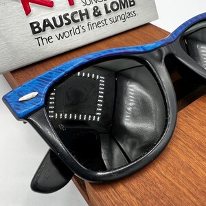 Op de afbeelding: Zwarte zonnebril met blauw gemarmerde armen. De zonnebril heeft donkere glazen en een zilveren accent op de slapen. De tekst "BAUSCH & LOMB The world's finest sunglass." is zichtbaar op de achtergrond.