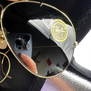 May include: Gold-rimmed sunglasses with dark lenses. The Ray-Ban logo is visible on the lens. The sunglasses are in a black case. The lenses reflect the surrounding environment.