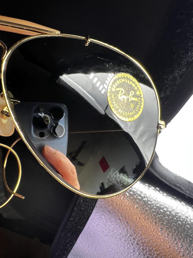 May include: Gold-rimmed sunglasses with dark lenses. The Ray-Ban logo is visible on the lens. The sunglasses are in a black case. The lenses reflect the surrounding environment.