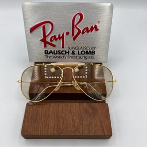 May include: Gold aviator sunglasses with brown lenses, resting on a wooden display stand. The stand has a silver plaque with the text "Ray-Ban Sunglasses by Bausch & Lomb The world's finest sunglass."