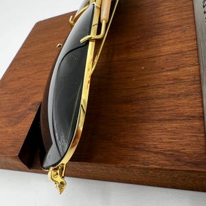 Pode incluir: &Oacute;culos de sol com arma&ccedil;&atilde;o dourada e lentes escuras. Os &oacute;culos s&atilde;o exibidos em um suporte de madeira. A arma&ccedil;&atilde;o tem um design decorativo perto das dobradi&ccedil;as. As lentes s&atilde;o escuras e refletivas.