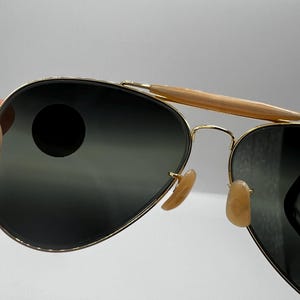 May include: Aviator sunglasses with gold-tone frames and dark lenses. The sunglasses feature a double bridge and light brown nose pads. The top bar is a light brown color. The lenses are a dark gray color.