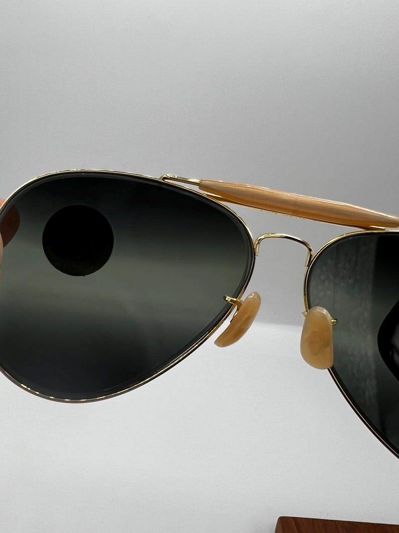 May include: Aviator sunglasses with gold-tone frames and dark lenses. The sunglasses feature a double bridge and light brown nose pads. The top bar is a light brown color. The lenses are a dark gray color.