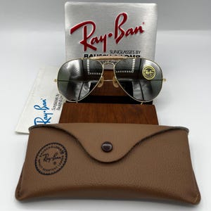 May include: Gold-framed Ray-Ban sunglasses with dark lenses, resting on a wooden display stand. A brown leather case and a silver sign with the Ray-Ban logo are also visible. The sunglasses have a classic aviator design.