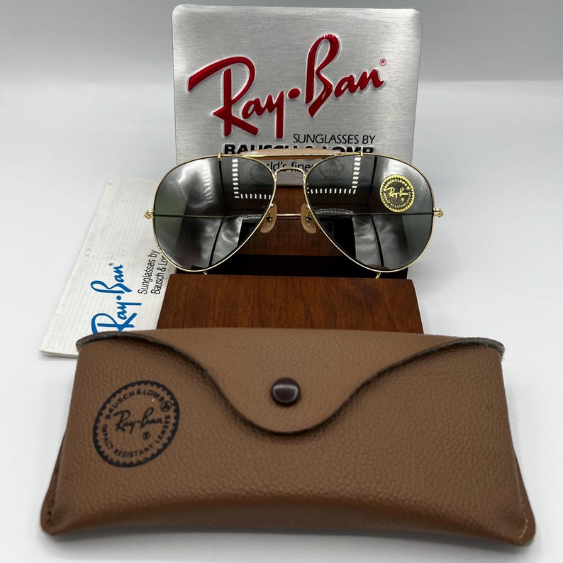 May include: Gold-framed Ray-Ban sunglasses with dark lenses, resting on a wooden display stand. A brown leather case and a silver sign with the Ray-Ban logo are also visible. The sunglasses have a classic aviator design.