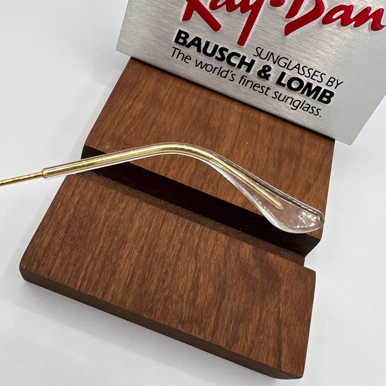 May include: A gold-colored metal temple tip for eyeglasses with a clear plastic end. The temple tip is resting on a wooden surface.